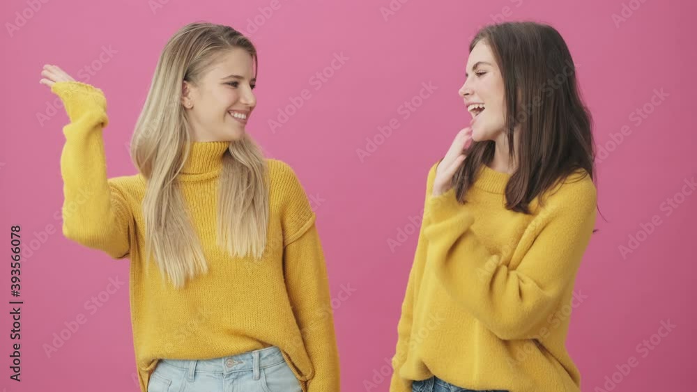 Two smiling women wearing yellow sweaters are giving five to each other standing isolated over pink background in studio Two smiling women wearing yellow sweaters are giving five to each other standing isolated over pink background in studio
