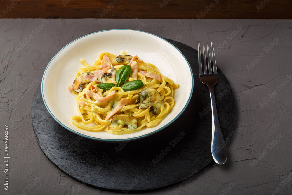a plate of pasta with ham and mushrooms in a creamy sauce with basil on a black round stand with a fork on a gray background. a plate of pasta with ham and mushrooms in a creamy sauce with basil on a black round stand with a fork on a gray background.