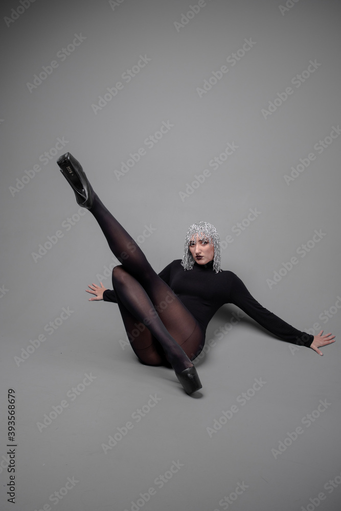 Obraz premium a long-legged girl in a silver wig in a black bodysuit and stockings in a photo studio in unusual poses on a gray background