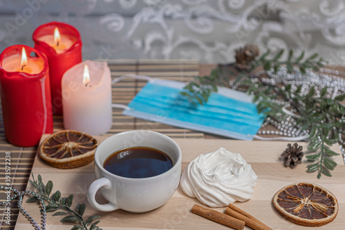 Medical mask, Christmas candles, coffee mug on a wooden table, air meringue, dried orange, cinnamon and accessories for winter holidays. New Year