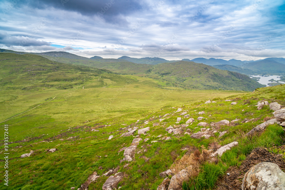 Panoramic view of the Irish countryside with trees, green vegetation and lakes with mountains and hills, cloudy day in Ireland. Girl sitting on the edge