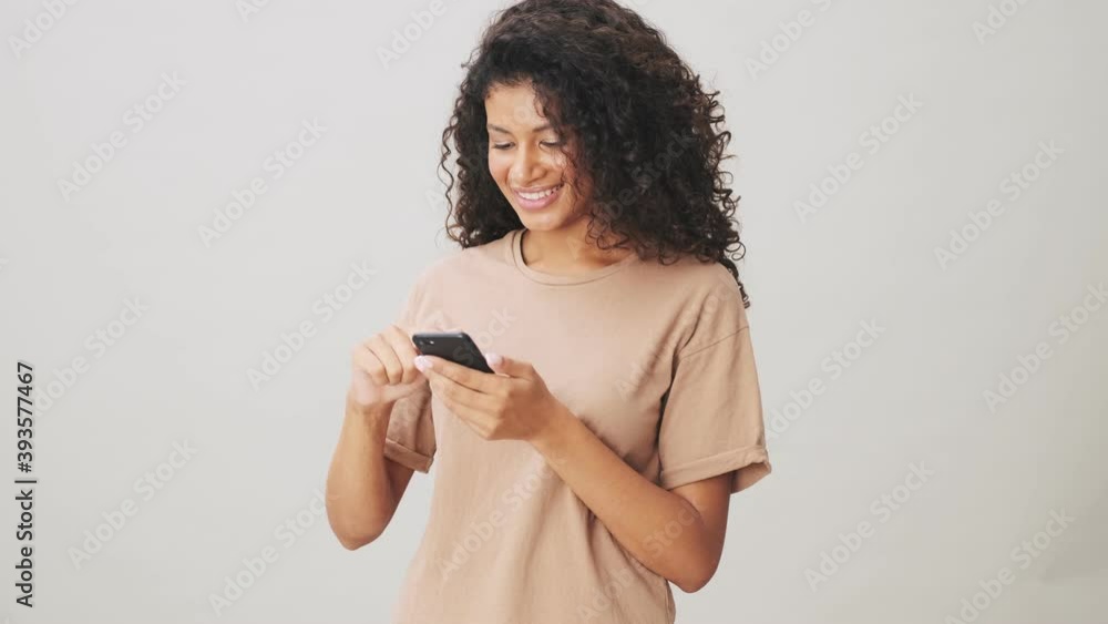 A smiling african american woman is swiping on her smartphone standing isolated over beige background A smiling african american woman is swiping on her smartphone standing isolated over beige background