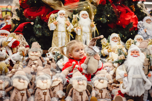 A little girl in a red suit sitting among New Year's toys on a shop window under the Christmas tree. Toy Santa Claus, St. Nicholas, gnomes and other characters look at an interesting girl with tails