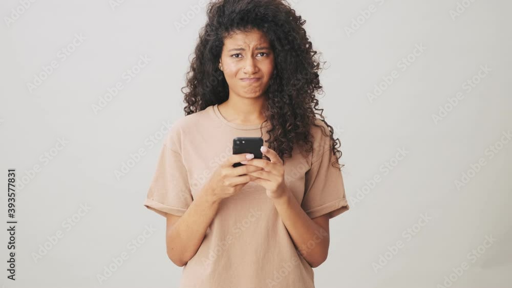 A sad african american woman is using her smartphone standing isolated over beige background A sad african american woman is using her smartphone standing isolated over beige background