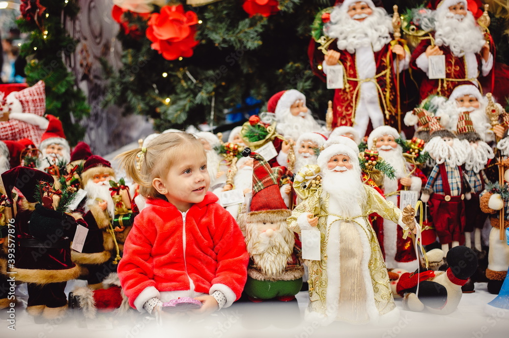 A little girl in a red suit sitting among New Year's toys on a shop window under the Christmas tree. Toy Santa Claus, gnomes. Christmas, New Year concept. Selective focus