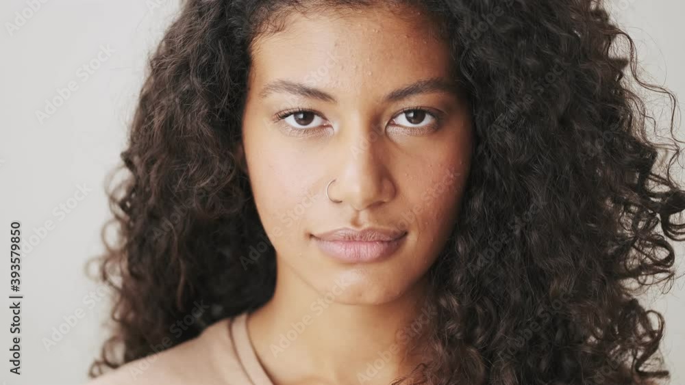 A close-up view of a charming african american woman is opening her eyes and looking to the camera standing isolated over beige background A close-up view of a charming african american woman is opening her eyes and looking to the camera standing isolated over beige background