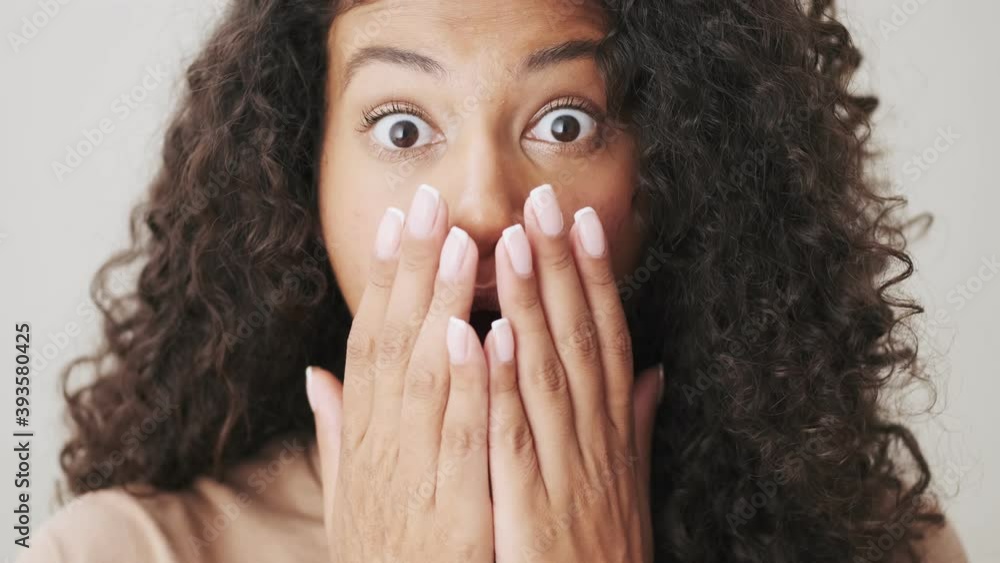 A close-up view of a surprised african american woman is looking to the camera standing isolated over beige background A close-up view of a surprised african american woman is looking to the camera standing isolated over beige background
