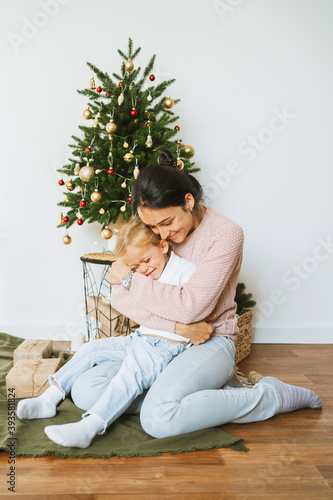 Happy young mom and little daughter cute hug under the Christmas tree in the room.Merry christmas and happy new year concept. Family cozy moments.Lifestyle. photo