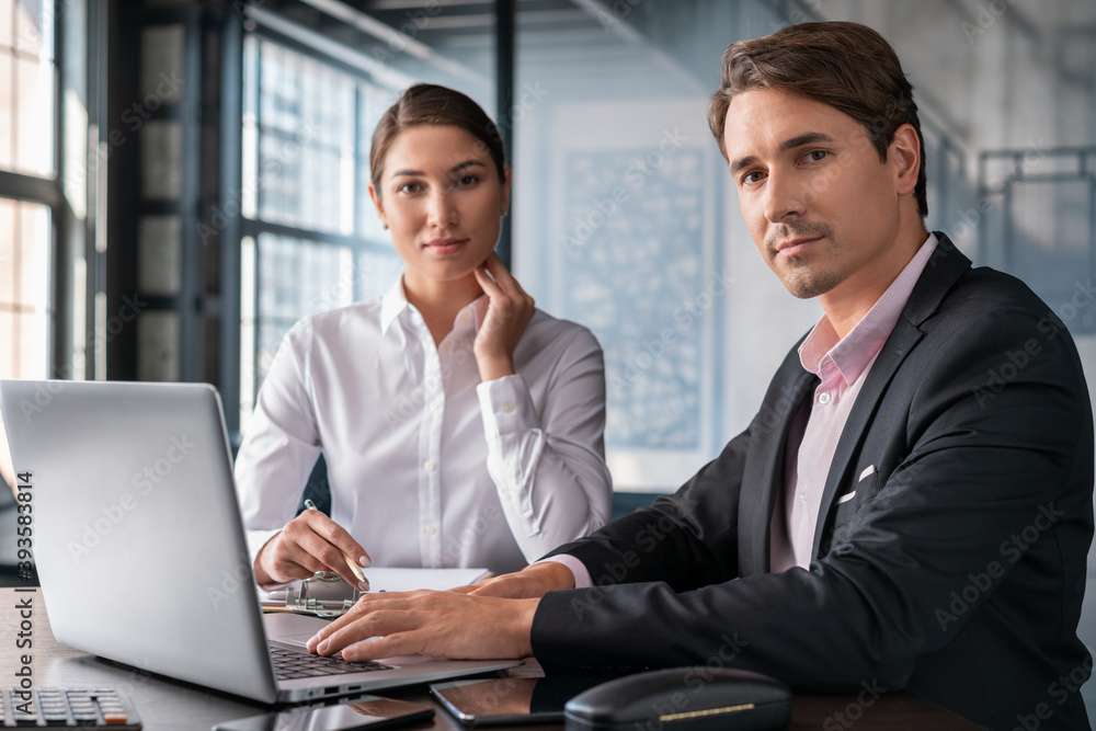Male and female office managers discussing work, man in black suit and woman in white shirt looking at the camera. Business partners in office room, concept of teamwork Male and female office managers discussing work, man in black suit and woman in white shirt looking at the camera. Business partners in office room, concept of teamwork