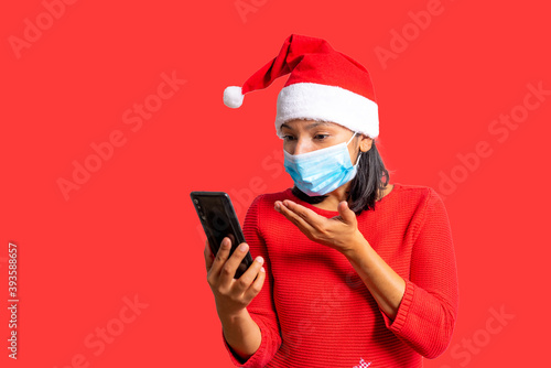 covid christmas concept. young woman with santa claus hat and face mask making mobile phone video call, concept of travel restrictions and lockdowns during holidays season. white background