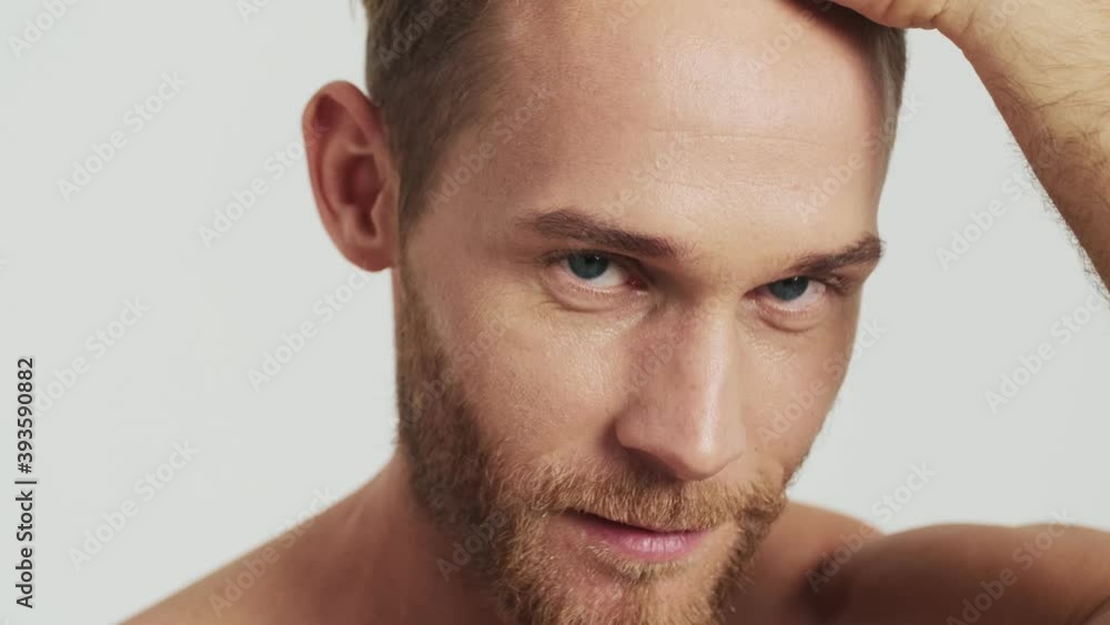 A close-up view of an attractive blonde man is touching his hair while looking to the camera standing isolated over a grey background in the studio A close-up view of an attractive blonde man is touching his hair while looking to the camera standing isolated over a grey background in the studio