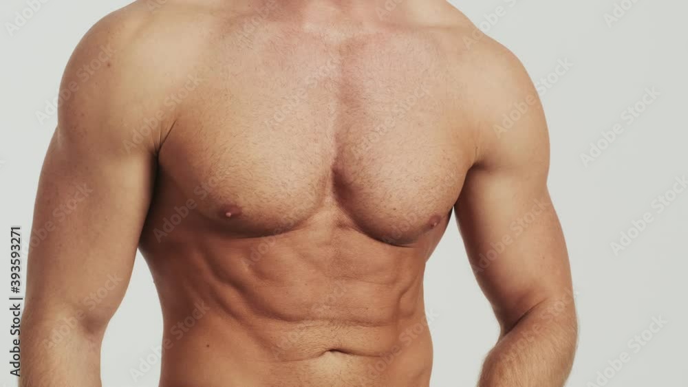 A cropped-view of a half-naked man is showing his press muscles posing to the camera standing isolated over a grey background in the studio A cropped-view of a half-naked man is showing his press muscles posing to the camera standing isolated over a grey background in the studio