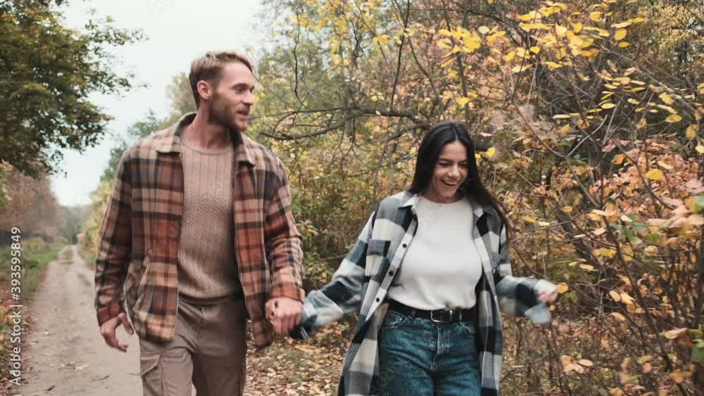 A smiling young couple in love man and woman are running while holding hands in the autumn park together A smiling young couple in love man and woman are running while holding hands in the autumn park together