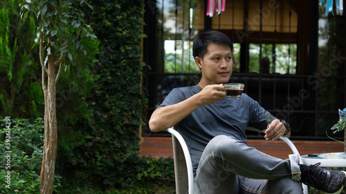 An adult Asian man wearing a gray shirt and jeans, sitting on a white shirt in the garden in front of the house, holding a cup of black coffee. © jakkrit