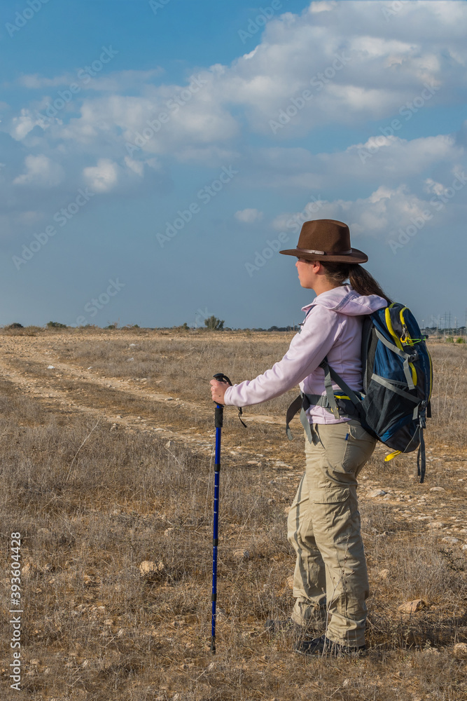 Young woman hiker with hipster hat, backpack, and tracking stick, walks through a field. Young woman hiker with hipster hat, backpack, and tracking stick, walks through a field.