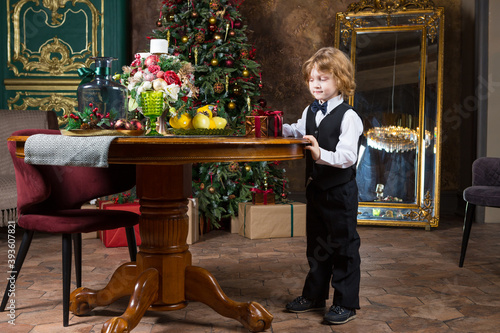 man-child wearing in black bow tie, whire shirt, black suit and shoes stands near round table in xmas decorated drawing-room with gift-box in his hands photo