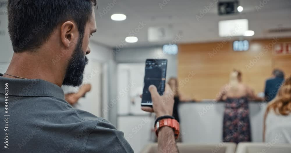 Young man using smartphone while sitting at hospital lobby waiting doctor visit. Man mobile technology user working in digital applications gadget searching information online, texting messages Young man using smartphone while sitting at hospital lobby waiting doctor visit. Man mobile technology user working in digital applications gadget searching information online, texting messages