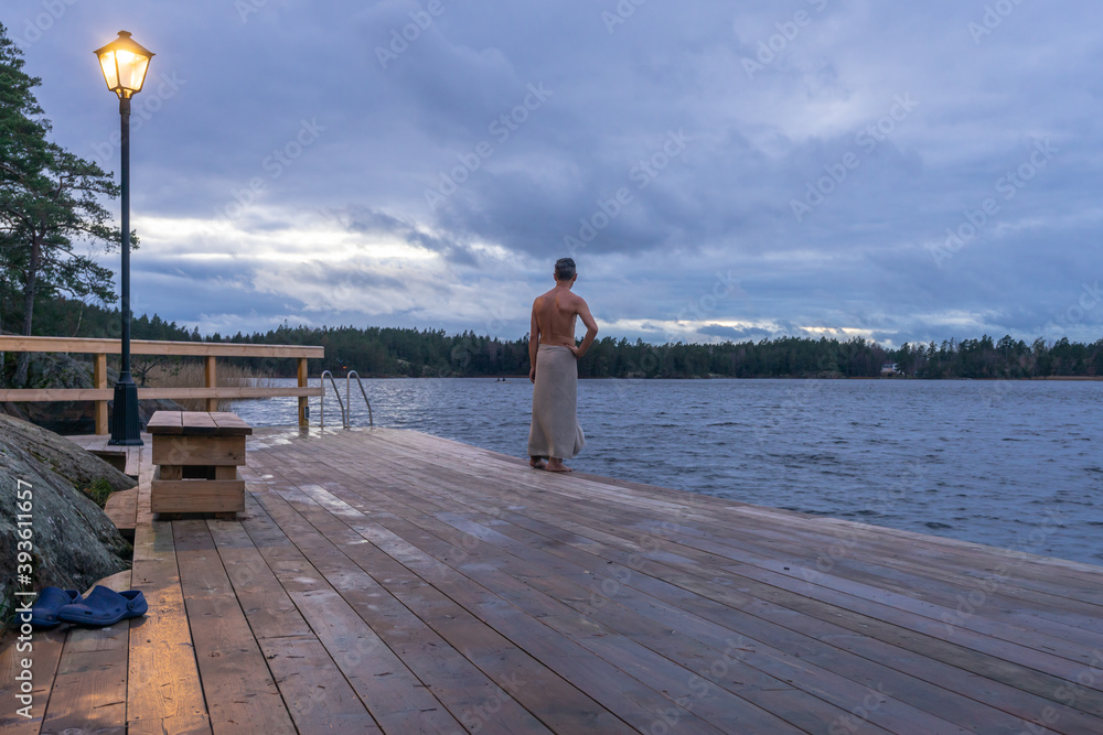 Finnish sauna. A man wrapped in a towel going for swimming in cold water. Wooden bathhouse or sauna with descent into the lake water. People enjoying hot bathing. Healthy lifestyle. Sunset on the lake