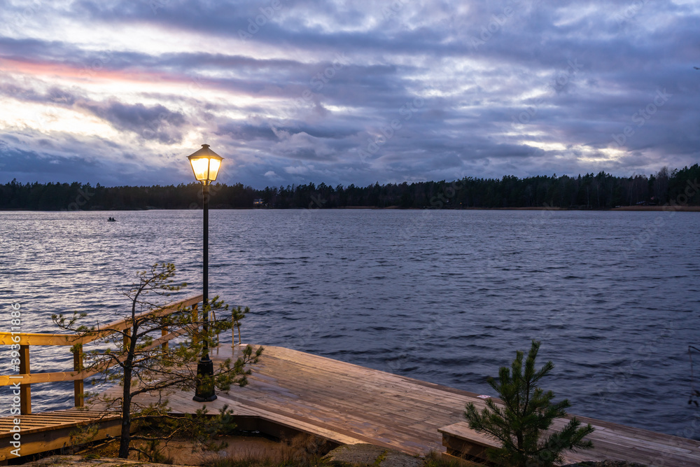 Obraz premium Finnish or russian sauna. Sunset on the lake in the evening. Wooden bath or sauna with descent into the water. Wooden flooring of planks on the shore for swimming in the cold water of the lake.