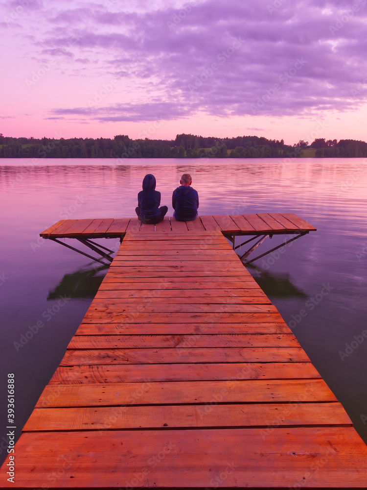 Two silhouettes of people contemplating nature and a water (lake) during a sunset on the end of a long jetty.