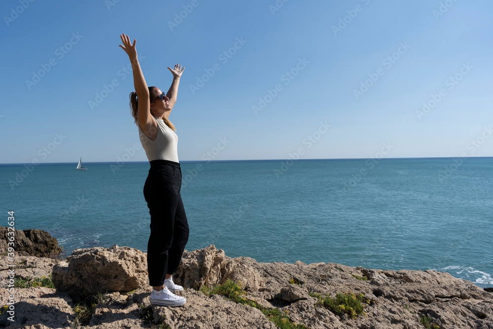 Pretty female celebrating and enjoying with raised up open arms standing on the rocks next to the sea.Freedom, happiness and people success lifestyle concept.Summer background.Copy space text. Pretty female celebrating and enjoying with raised up open arms standing on the rocks next to the sea.Freedom, happiness and people success lifestyle concept.Summer background.Copy space text.