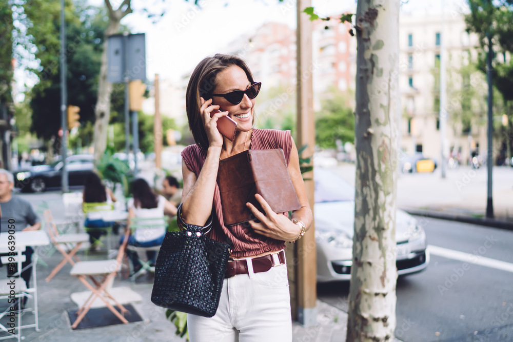 Fototapeta premium Prosperous caucasian businesswoman with notepad having working conversation on mobile phone walking on city street, successful pretty female in sunglasses talking on smartphone via roaming tariffs
