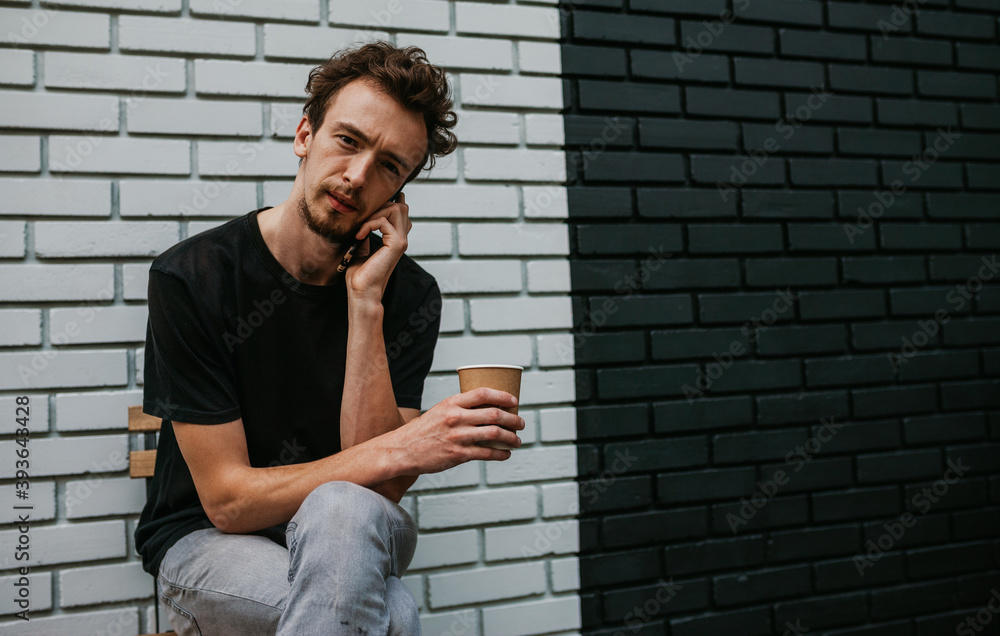 A young charismatic guy, a hipster, drinks coffee and speaks on the phone against the background of a brick wall. Dressed in a black T-shirt. Mock-up for printing A young charismatic guy, a hipster, drinks coffee and speaks on the phone against the background of a brick wall. Dressed in a black T-shirt. Mock-up for printing