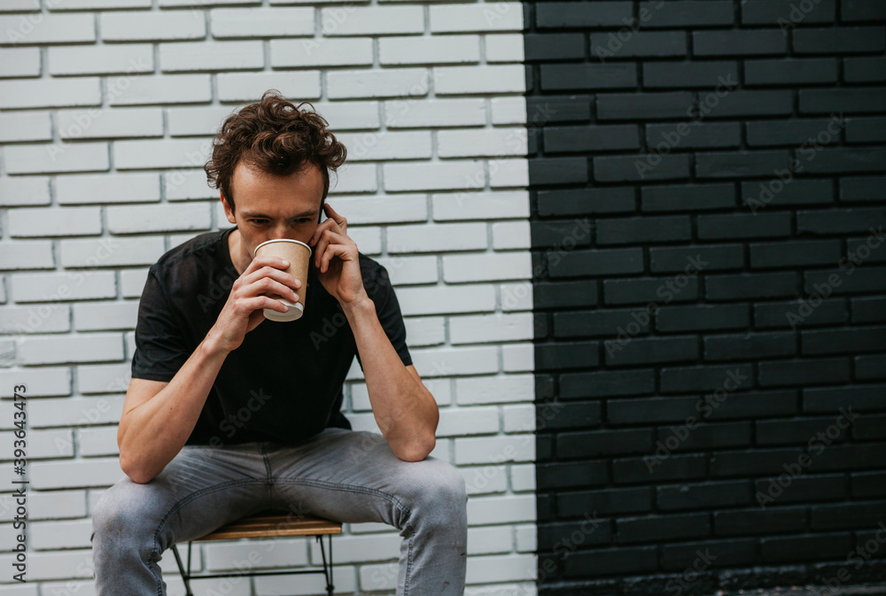 A young charismatic guy, a hipster, drinks coffee and speaks on the phone against the background of a brick wall. Dressed in a black T-shirt. Mock-up for printing A young charismatic guy, a hipster, drinks coffee and speaks on the phone against the background of a brick wall. Dressed in a black T-shirt. Mock-up for printing