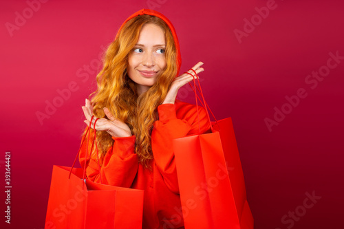 Redhead girl with freckles, pensive look holds red gift bags on red background.