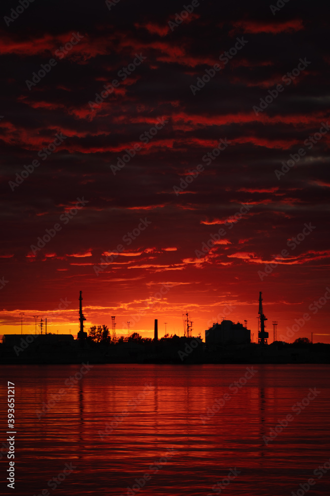 Bloody sunset with black industrial city  silhouette. Beautiful and scary dark red sundown on the beach with old factory buildings. Cloudy summer sky in the evening. Vertical landscape