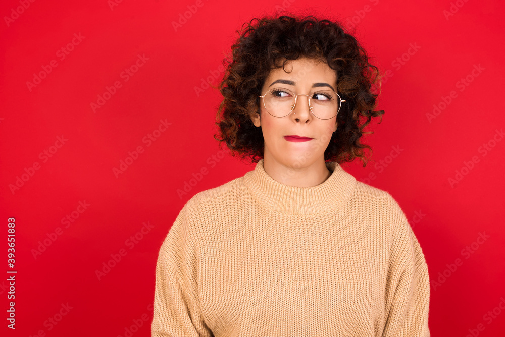 Amazed puzzled Young beautiful Arab woman wearing beige sweater against red background, curves lips and has worried look, sees something awful in front. Amazed puzzled Young beautiful Arab woman wearing beige sweater against red background, curves lips and has worried look, sees something awful in front.