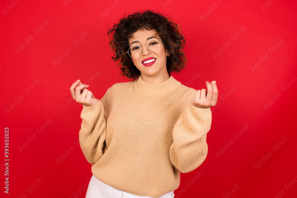 Young beautiful Arab woman wearing beige sweater against red background, making money gesture. Young beautiful Arab woman wearing beige sweater against red background, making money gesture.
