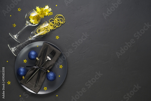 Festive table setting with black plate, cutlery, wine glasses with gold confetti and blue xmas balls on dark tabletop. Christmas table setting. Flat lay, top view. Minimal style, copy space