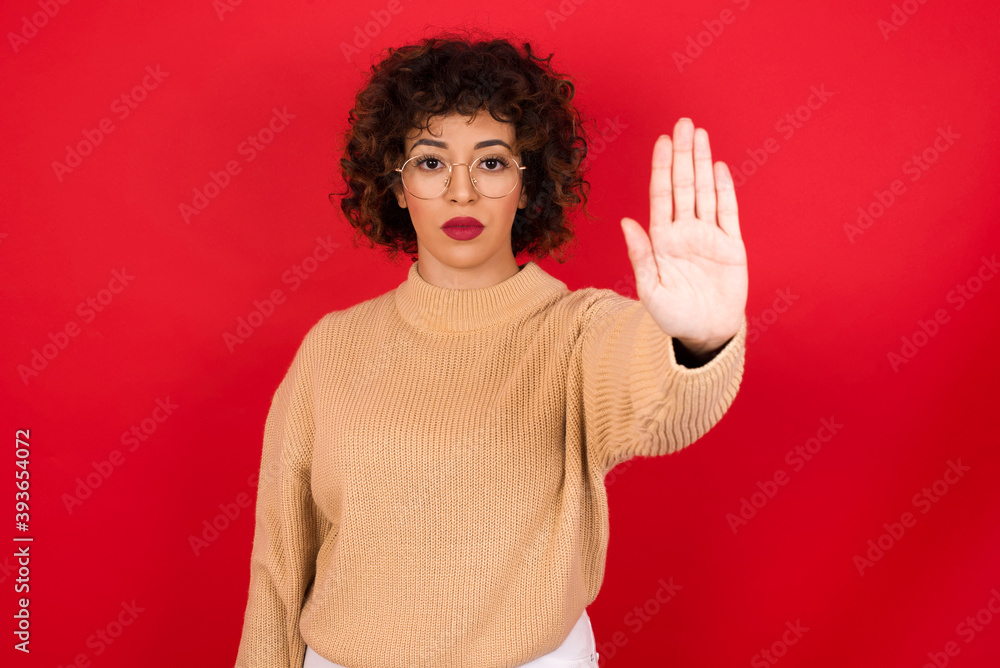 Young beautiful Arab woman wearing beige sweater against red background doing stop gesture with palm of the hand. Warning expression with negative and serious gesture on the face. Young beautiful Arab woman wearing beige sweater against red background doing stop gesture with palm of the hand. Warning expression with negative and serious gesture on the face.