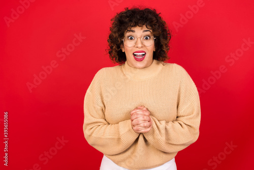 Happy Young beautiful Arab woman wearing beige sweater against red background stands against orange studio wall keeps hands on heart, swears be loyal, expresses gratitude. Honesty concept.