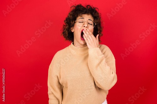 Sleepy Young beautiful Arab woman wearing beige sweater against red background yawning with messy hair, feeling tired after sleepless night, yawning, covering mouth with palm.