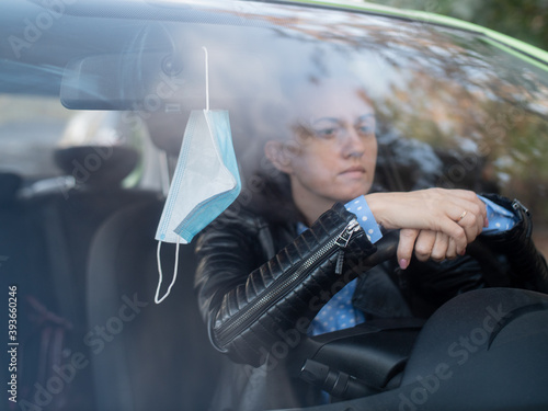 Sad woman driver sits in the car and holds his hands on the steering wheel of the car. Medical mask hanging on the mirror in the car