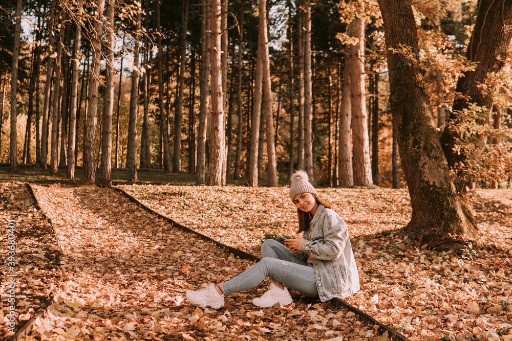 Fototapeta premium A smiling cute brunette caucasian girl with a hat, dressed in jeans sits in a park on the ground and uses the phone. Autumn walk in nature