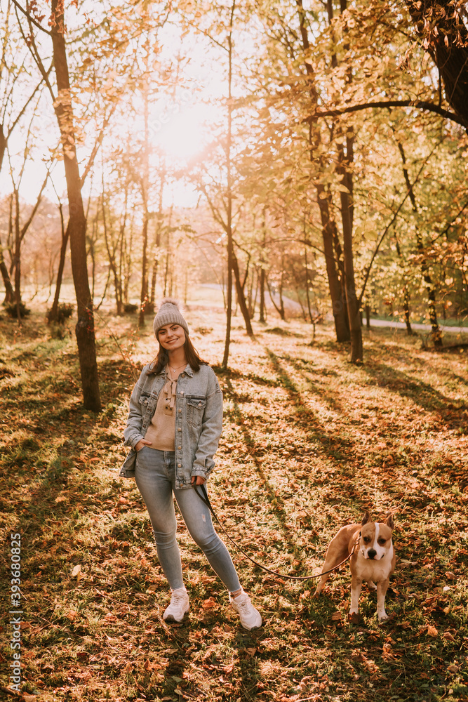 Fototapeta premium A young urban teenage girl with a hat in a denim coat standing in the park next to a brown American American Staffordshire Terrier. Autumn walk in nature
