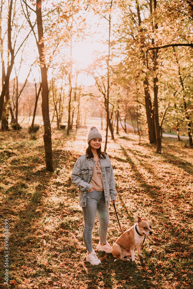 Full length of a young cute teenage girl standing in the park and holding her brown dog breed American Staffordshire Terrier. Autumn day in the woods