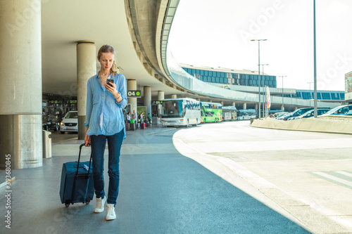 Beautiful modern woman tourist walking with a travel bag and waiting for a taxi with a smartphone in her hand on background of the airport building and transport stops on a sunny day.