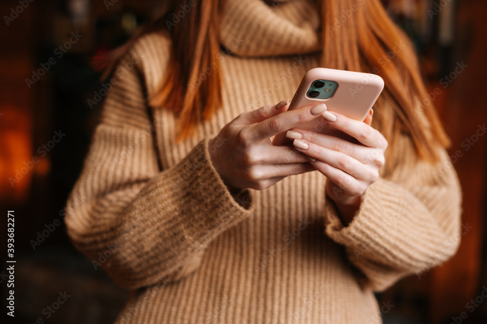 Close-up view of unrecognizable redhead woman wearing white winter sweater typing online message on mobile phone. Female with mobile phone spending Christmas time at home. Close-up view of unrecognizable redhead woman wearing white winter sweater typing online message on mobile phone. Female with mobile phone spending Christmas time at home.