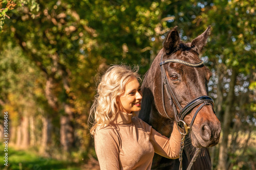 Front view, portrait. Female in casual clothes stands in the forest with her horse. In the autumn sun