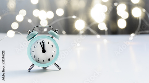 A small blue alarm clock shows 12 o'clock, stands on a light table with a blurred background and bokeh lights. Christmas and New Year concept.