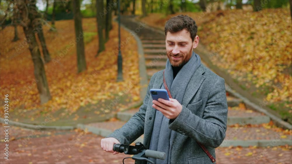 Smiling young stylish man in autumn park use phone stand with electric scooter. Ecological transportation. Feel happy. Urban outdoors. Slow motion Smiling young stylish man in autumn park use phone stand with electric scooter. Ecological transportation. Feel happy. Urban outdoors. Slow motion