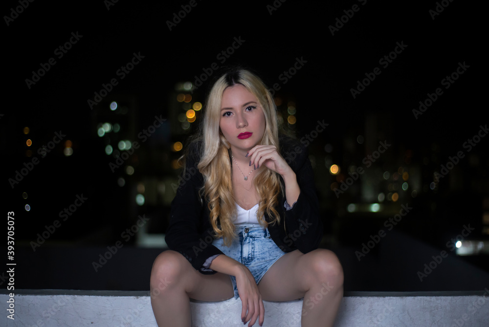 blonde girl with black jacket and shorts sit on a rooftop with blurry lights background blonde girl with black jacket and shorts sit on a rooftop with blurry lights background