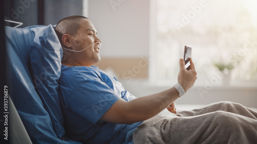 Medical Hospital Ward: Portrait of Handsome Chinese Man with Nasal Cannula Resting in Bed, Uses Smartphone to Video Chat with His Family, Friends, Shooting Blog. Surgery Recovery Room. Side View.
