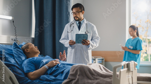 Hospital Ward  Friendly Chinese Doctor Talks to Handsome Male Patient Resting in Bed  Uses Tablet Computer  Explains Test Results. Physician Talks to Man Recovering after Successful Surgery