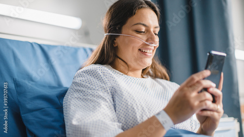 Medical Hospital Ward: Portrait of Beautiful Latin Woman with Nasal Cannula Resting in Bed, Uses Smartphone to Messaging with Her Family, Friends, Read News. Recovering after Successful Surgery.