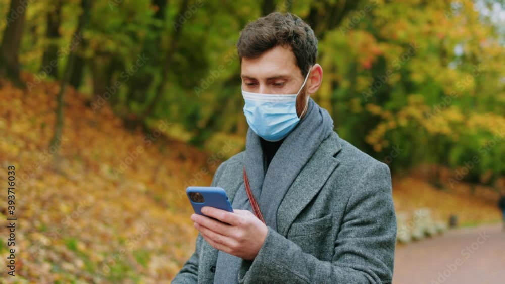 Stylish man with medical mask in autumn park use phone stand with electric scooter. Ecological transportation. Pandemic coronavirus. Urban outdoors. Slow motion Stylish man with medical mask in autumn park use phone stand with electric scooter. Ecological transportation. Pandemic coronavirus. Urban outdoors. Slow motion
